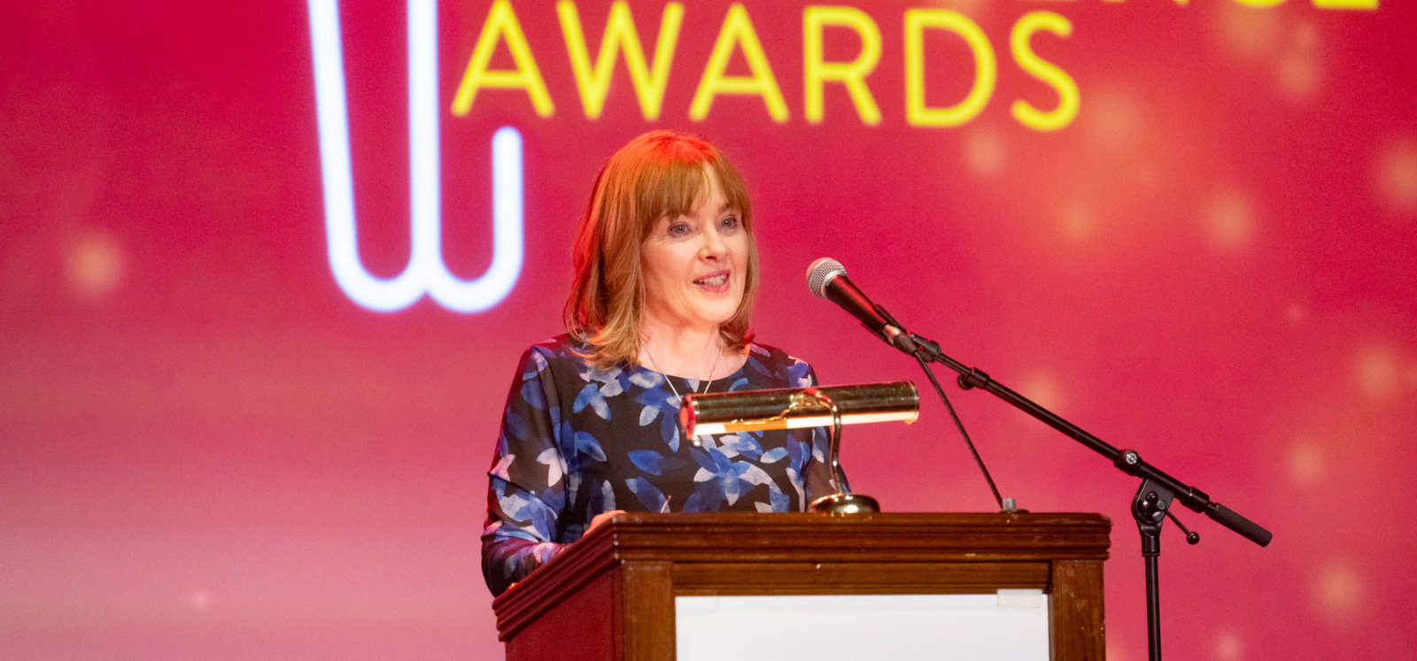 Brunette lady standing in front of a brown podium in front of red Staff Excellence Awards backdrop